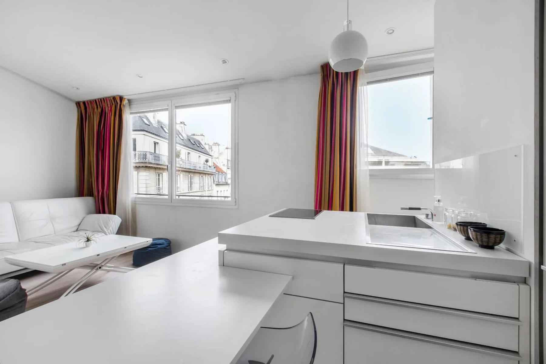 Una cocina luminosa con encimeras blancas, gabinetes y ventanas con vistas a edificios cercanos.