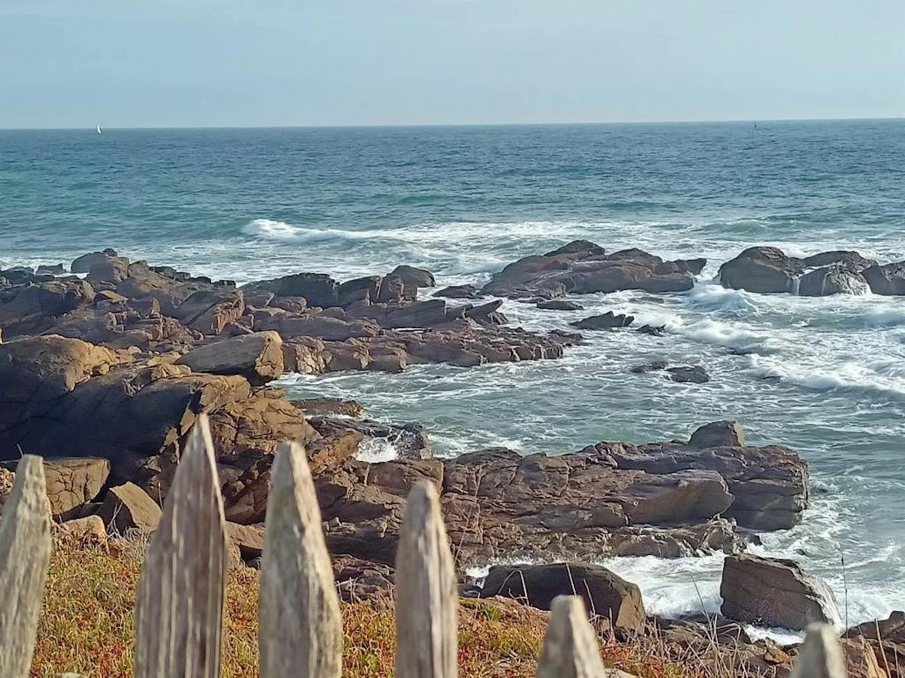 Ocean waves wash over a rocky shoreline beside a weathered wooden fence.