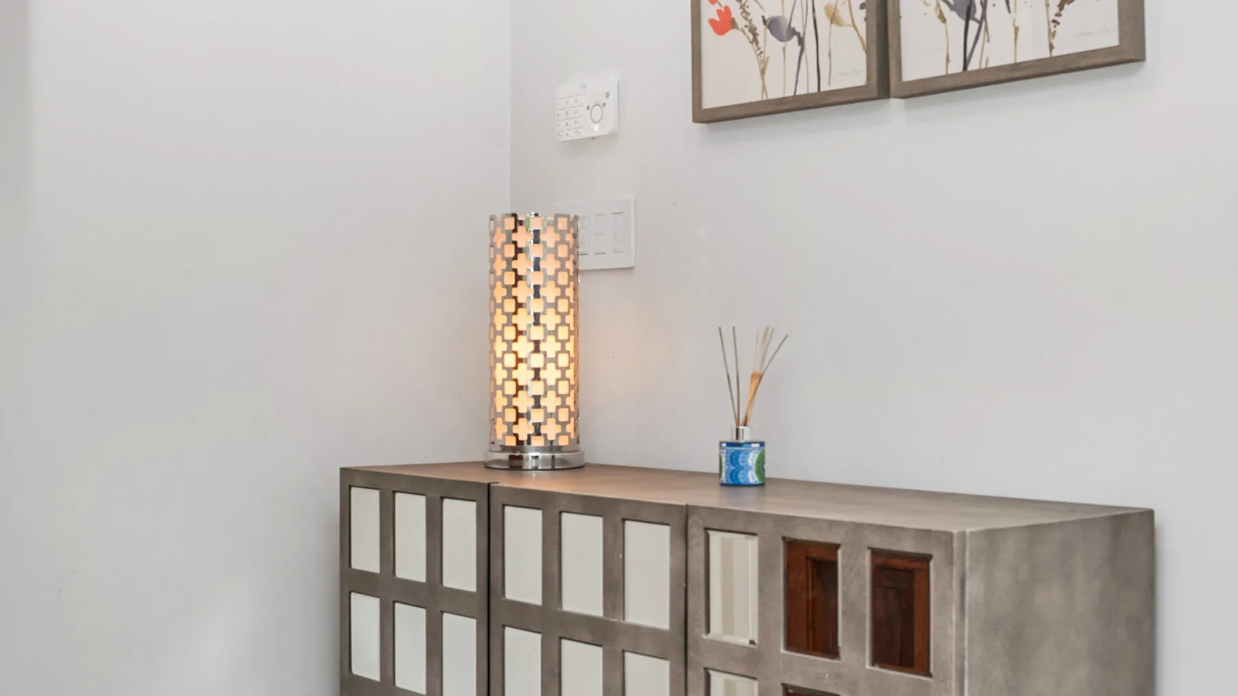 Una consola con una lámpara encendida y cuadros florales enmarcados junto a una pared blanca.