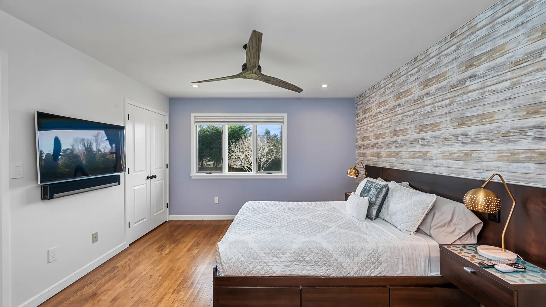 Un dormitorio con una cama grande, un televisor montado en la pared y una ventana con vista a los árboles.
