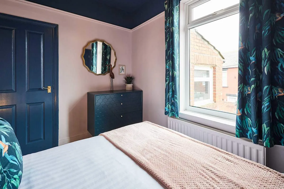 Un dormitorio con una cama, una cómoda azul oscuro y una ventana frente a edificios cercanos.