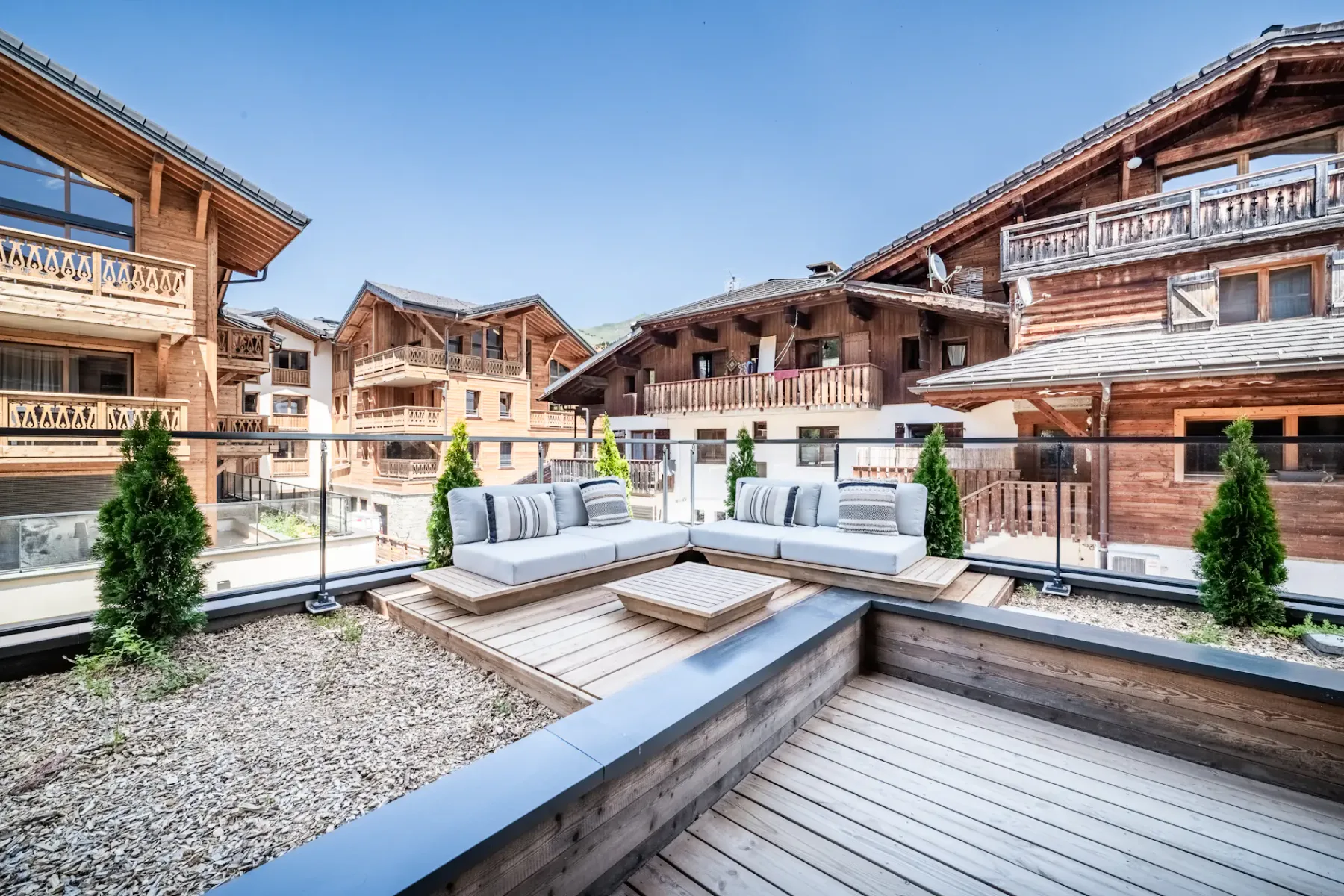 Outdoor wooden terrace with a table and cushioned chairs, overlooking an alpine-style village with wooden buildings under a clear blue sky.