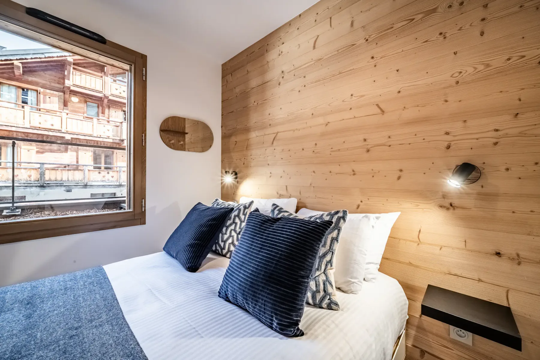 A cozy bedroom with a double bed, blue pillows, and a blanket. Wood-paneled wall with small light fixtures, a window, and clothes rack with hangers.