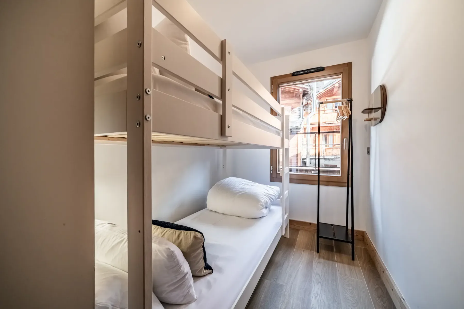 Small room with wooden bunk beds, white bedding, a window showing an outside view, and a black metal shelf against a white wall.