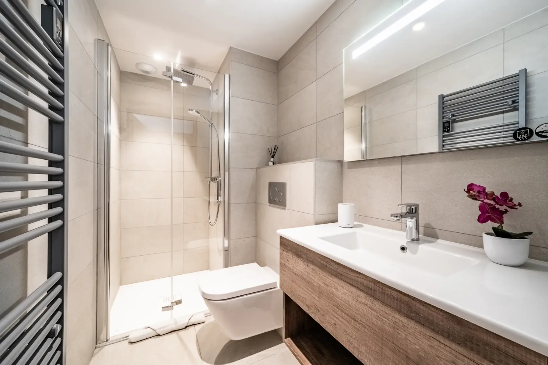 Modern bathroom with a glass shower, wall-mounted toilet, wooden vanity with sink, and illuminated mirror. A small potted orchid sits on the countertop.