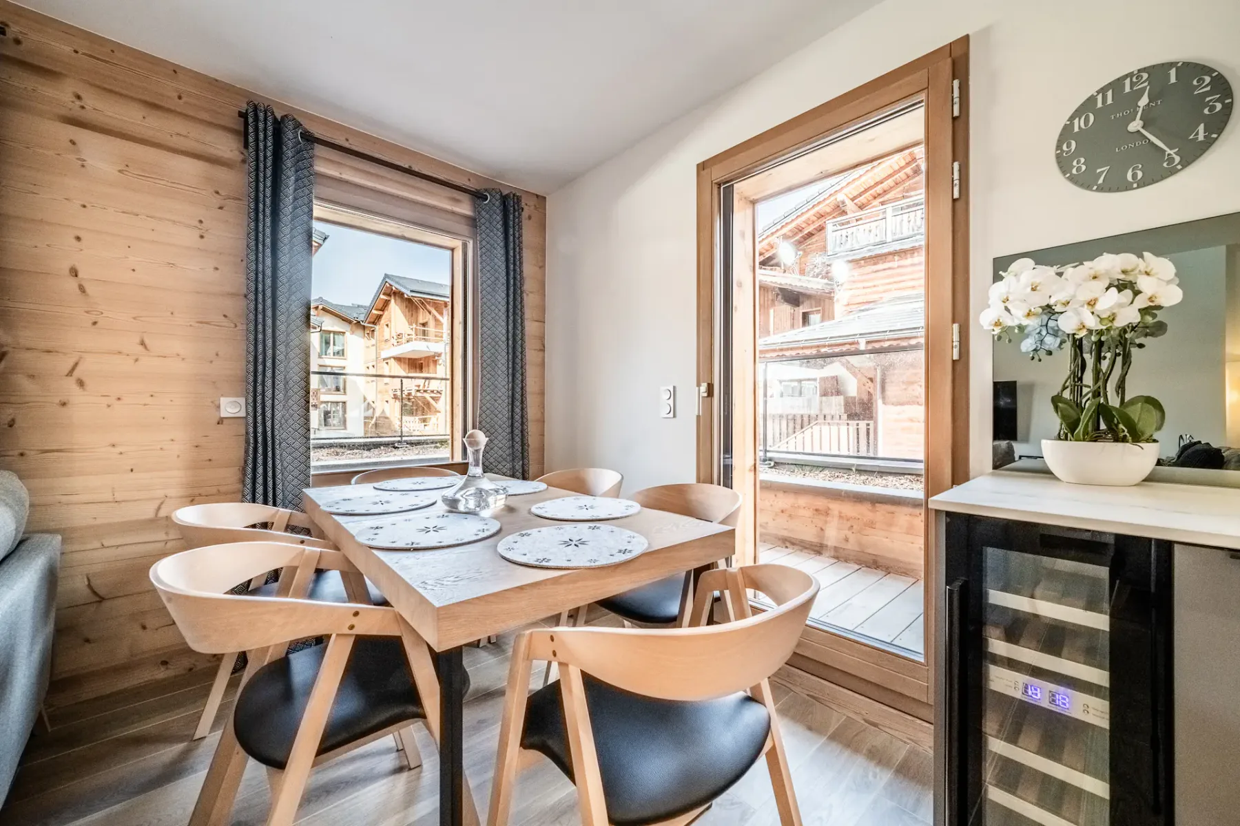 Modern kitchen and dining area with gray cabinetry, wooden floors, a small dining table with chairs, a sofa, and large windows with views of wooden buildings outside.