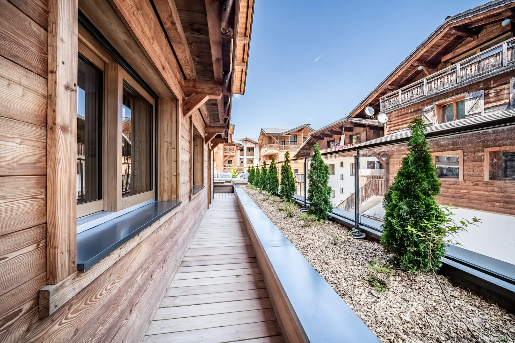 Outdoor wooden terrace with a table and cushioned chairs, overlooking an alpine-style village with wooden buildings under a clear blue sky.