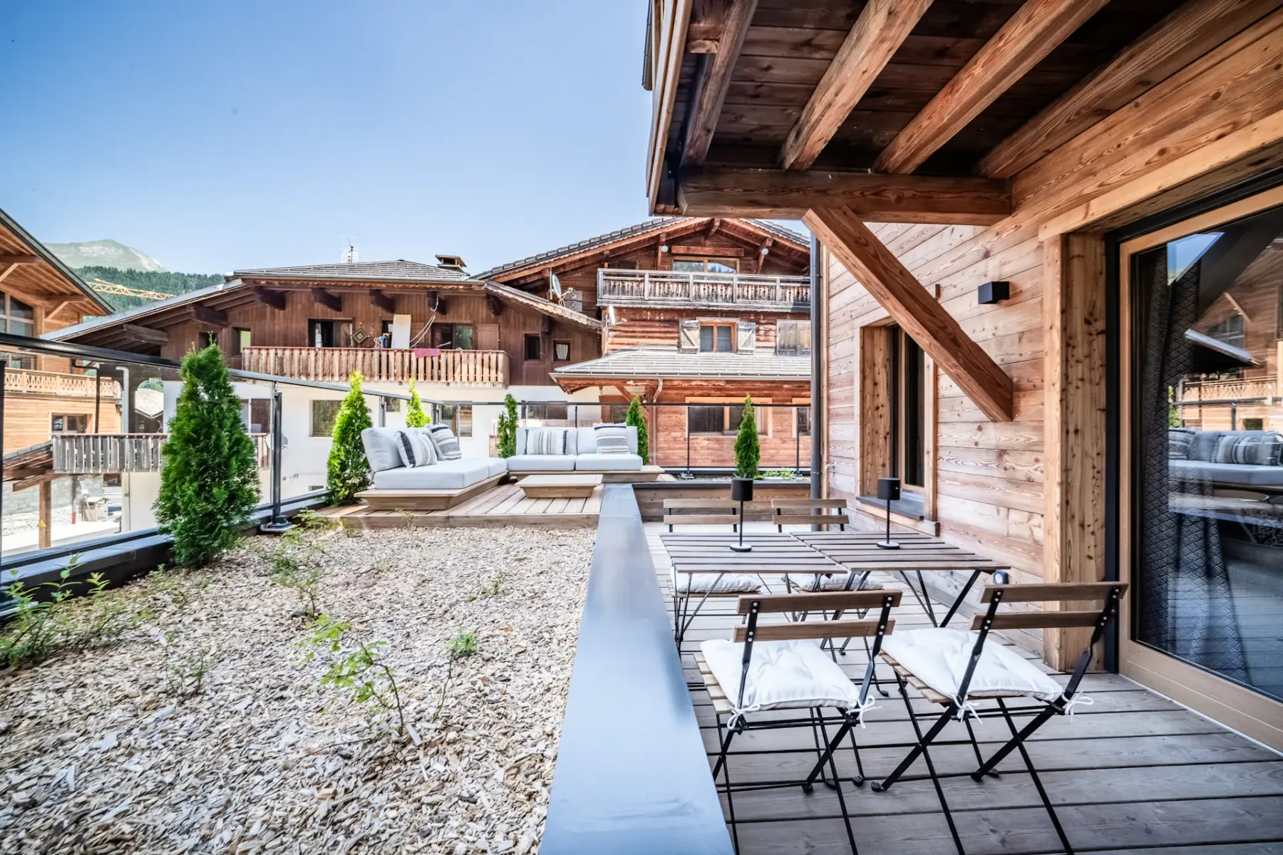 Outdoor wooden terrace with a table and cushioned chairs, overlooking an alpine-style village with wooden buildings under a clear blue sky.