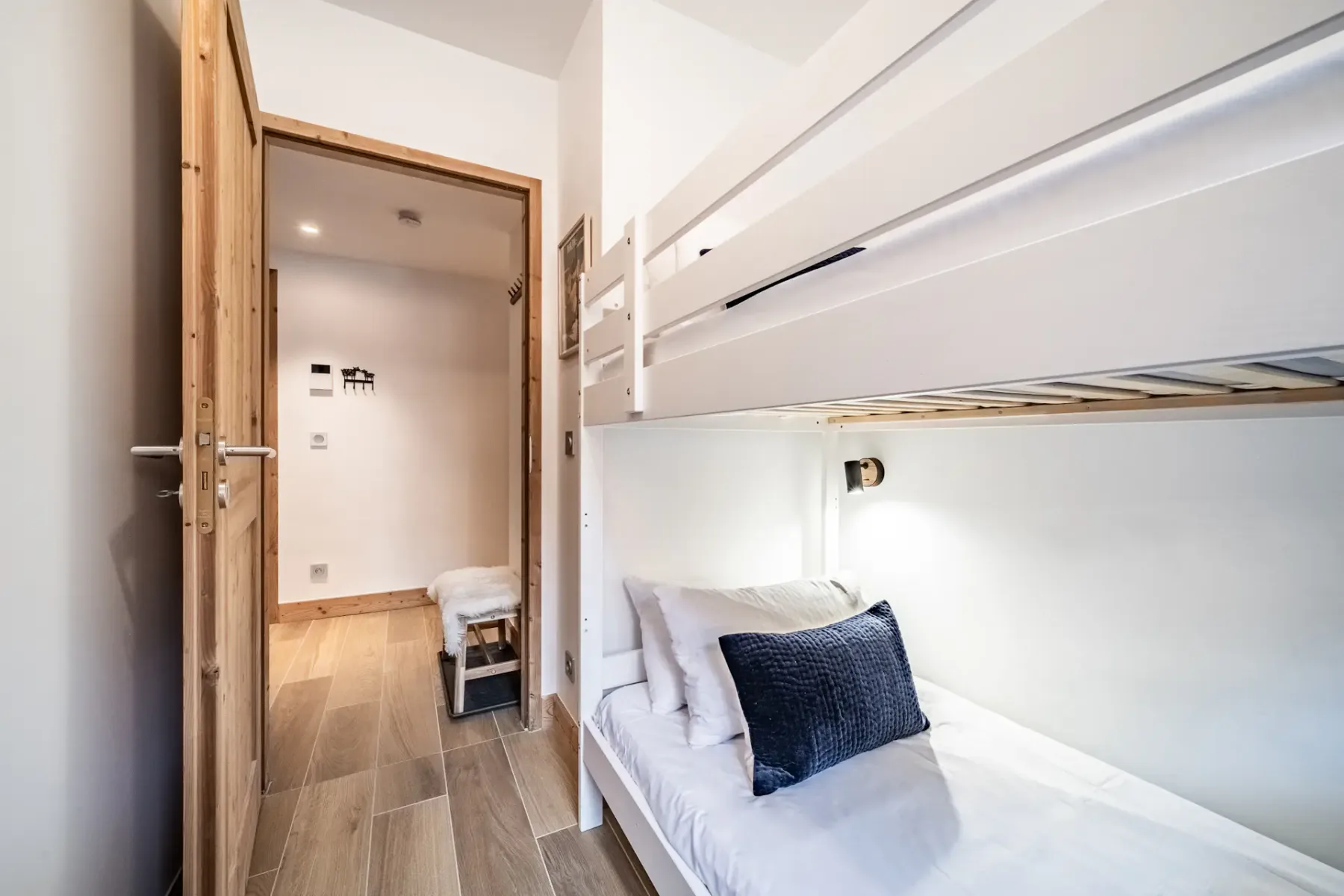 Small room with wooden bunk beds, white bedding, a window showing an outside view, and a black metal shelf against a white wall.