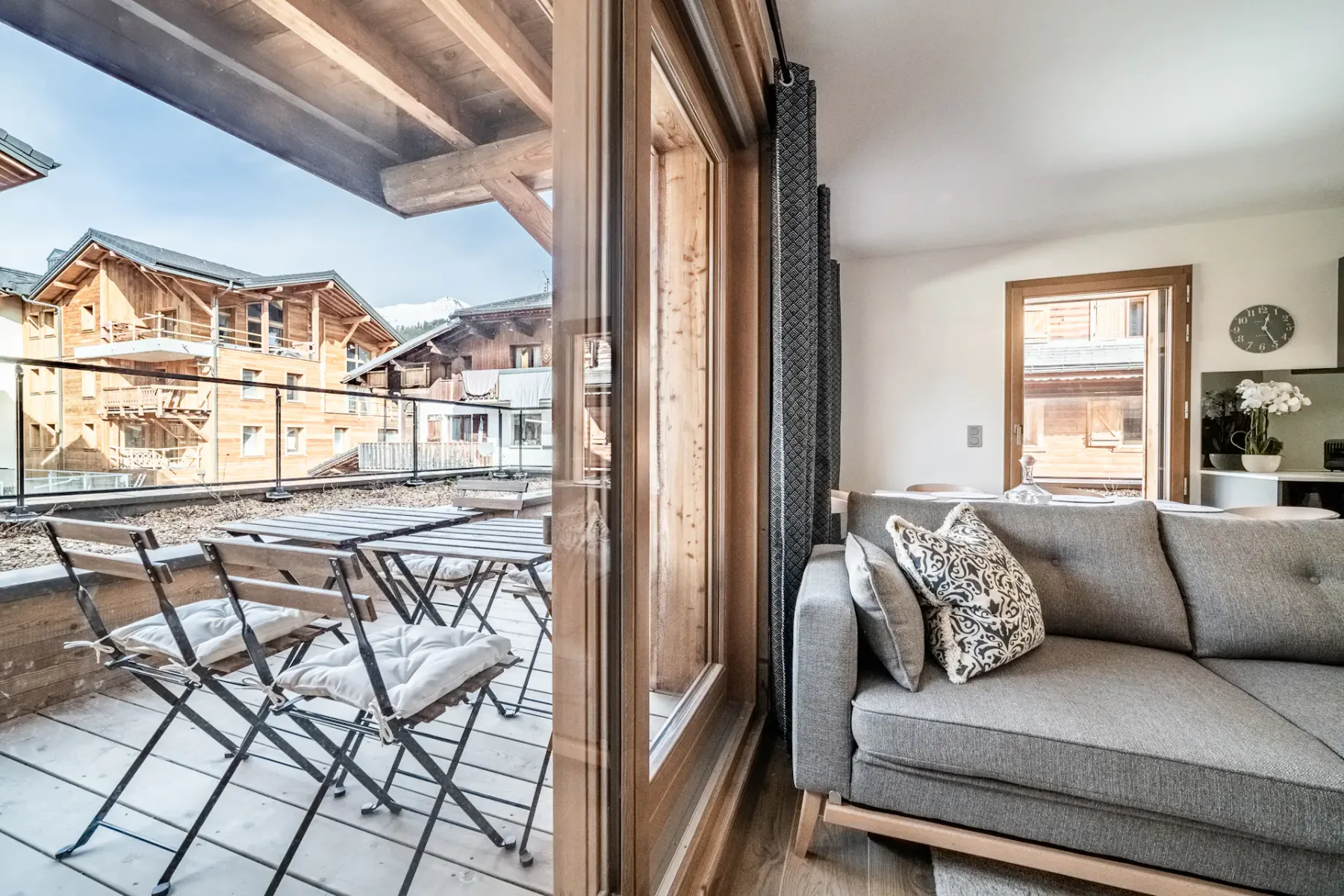 Modern living room with two beige sofas, a wooden coffee table, floor lamp, and wall-mounted TV. Glass doors open to a balcony with a view of neighboring buildings. Wood flooring and decor.