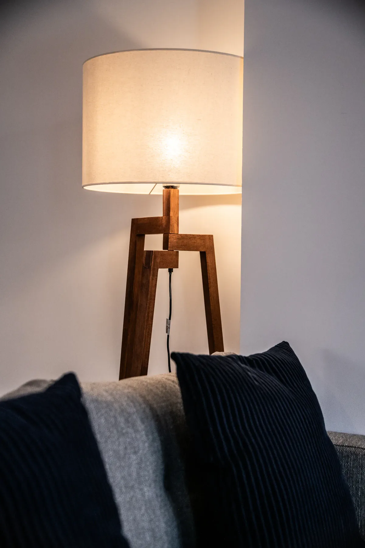 A wooden tripod floor lamp with a cylindrical white shade stands next to a sofa, which has two dark blue cushions.