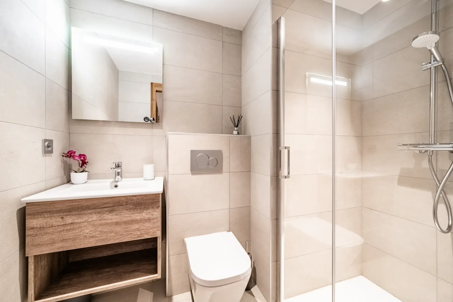 Modern bathroom with beige tiles, a glass shower enclosure, wall-mounted toilet, and wooden vanity with a rectangular sink and mirror. Pink flowers and a reed diffuser decorate the vanity.