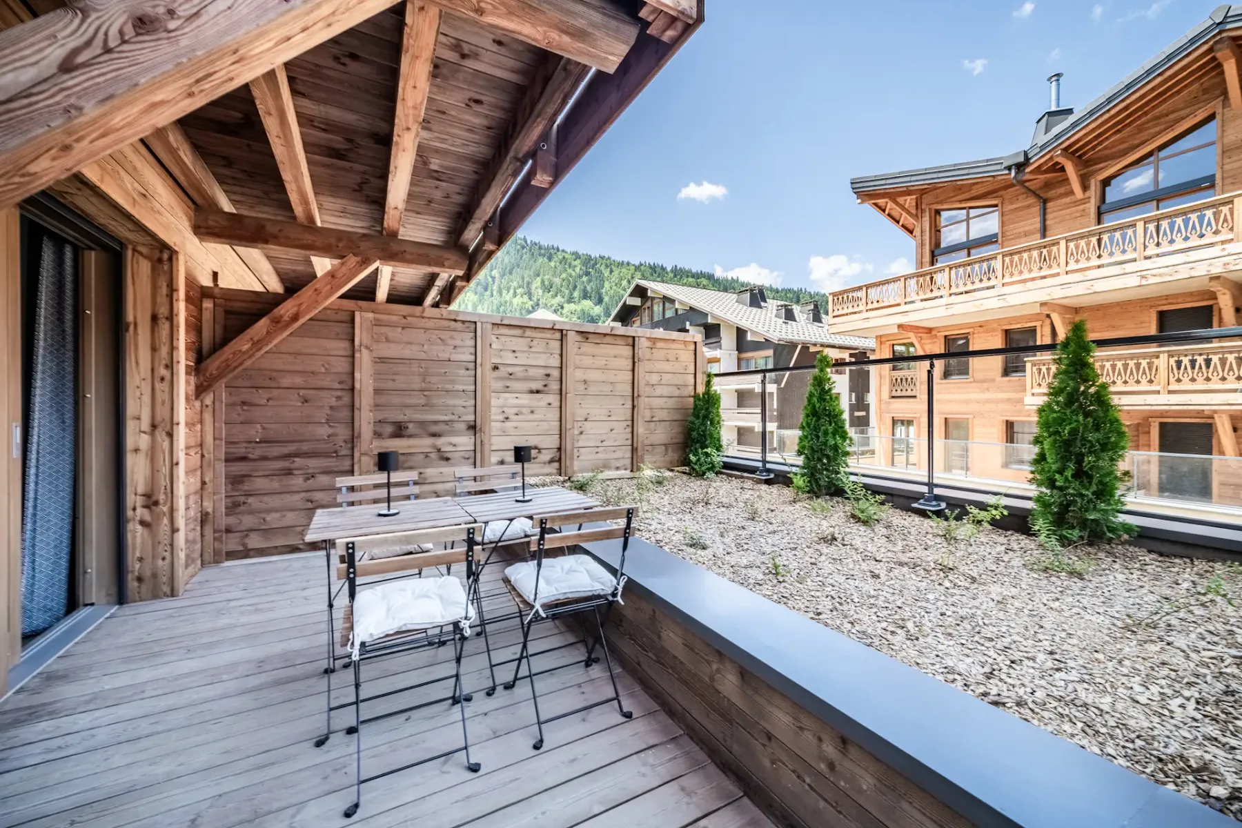 Outdoor wooden terrace with a table and cushioned chairs, overlooking an alpine-style village with wooden buildings under a clear blue sky.