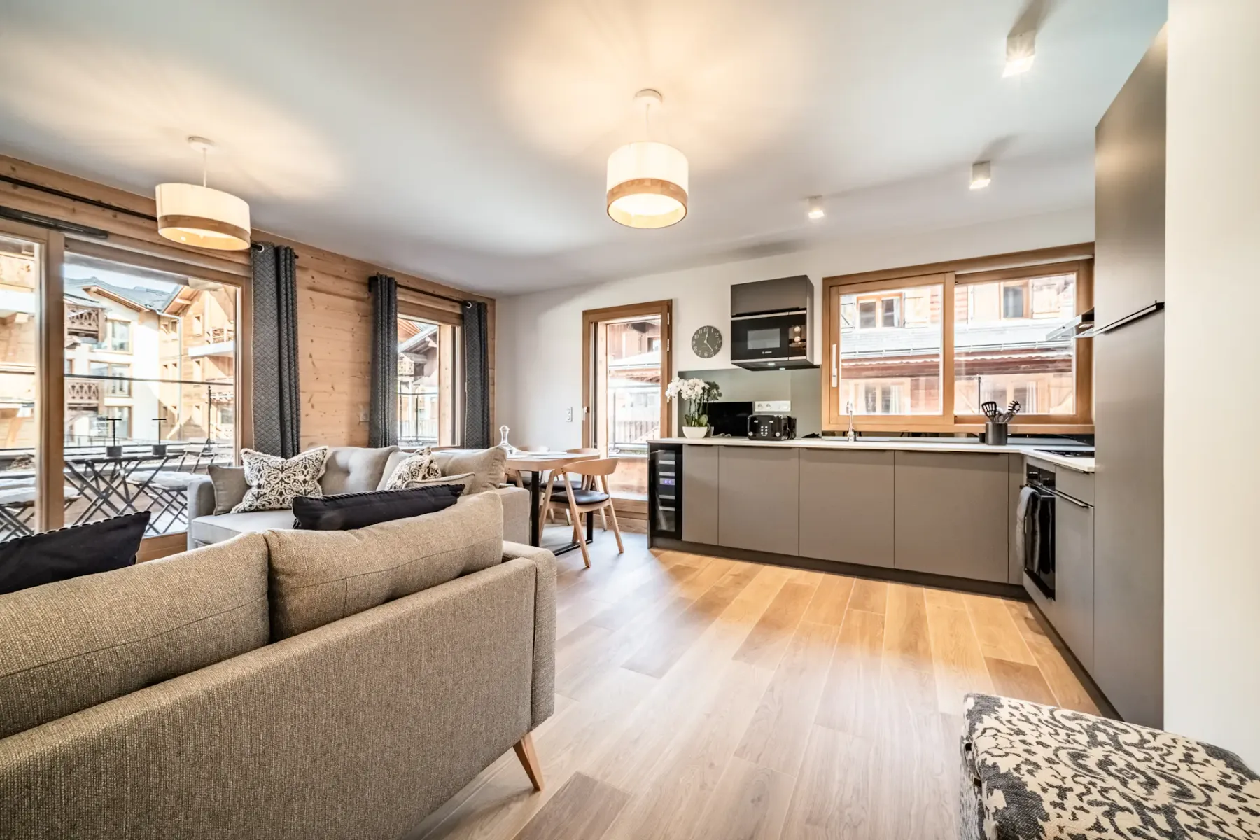 Modern kitchen and dining area with gray cabinetry, wooden floors, a small dining table with chairs, a sofa, and large windows with views of wooden buildings outside.