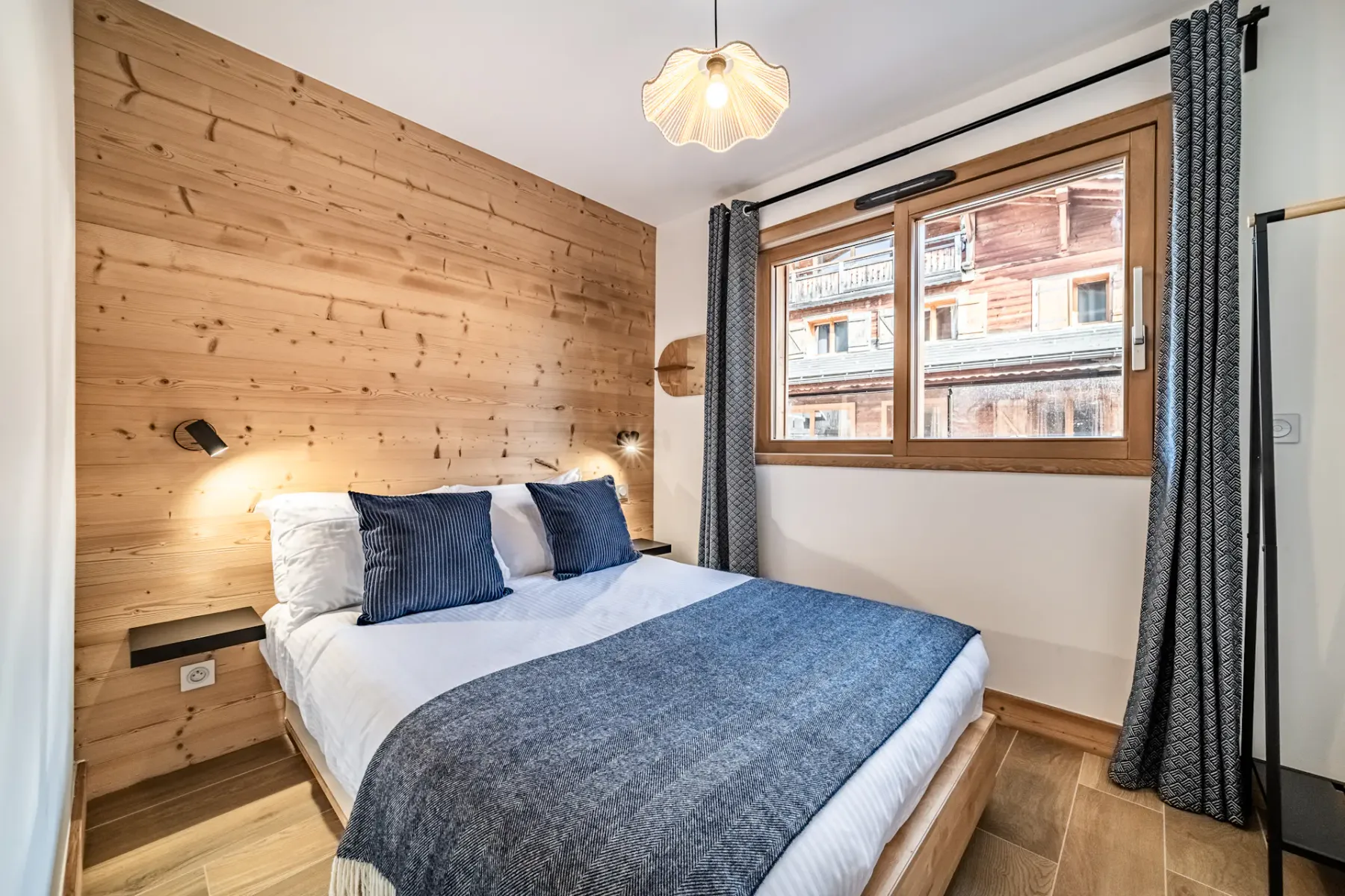 A cozy bedroom with wooden walls, featuring a double bed with blue pillows and a gray blanket. A window with curtains lets in natural light, and a pendant lamp hangs from the ceiling.