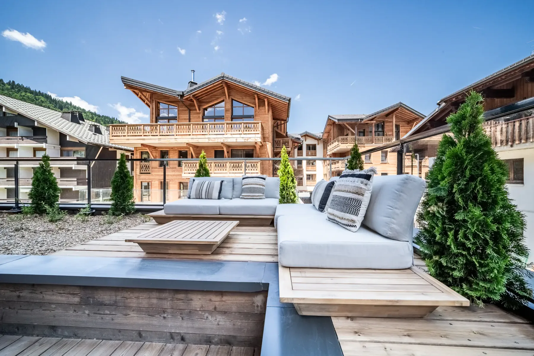 Outdoor wooden terrace with a table and cushioned chairs, overlooking an alpine-style village with wooden buildings under a clear blue sky.