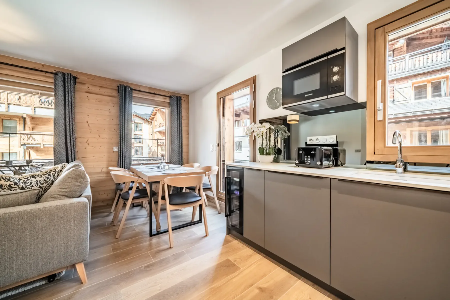 Modern kitchen and dining area with gray cabinetry, wooden floors, a small dining table with chairs, a sofa, and large windows with views of wooden buildings outside.