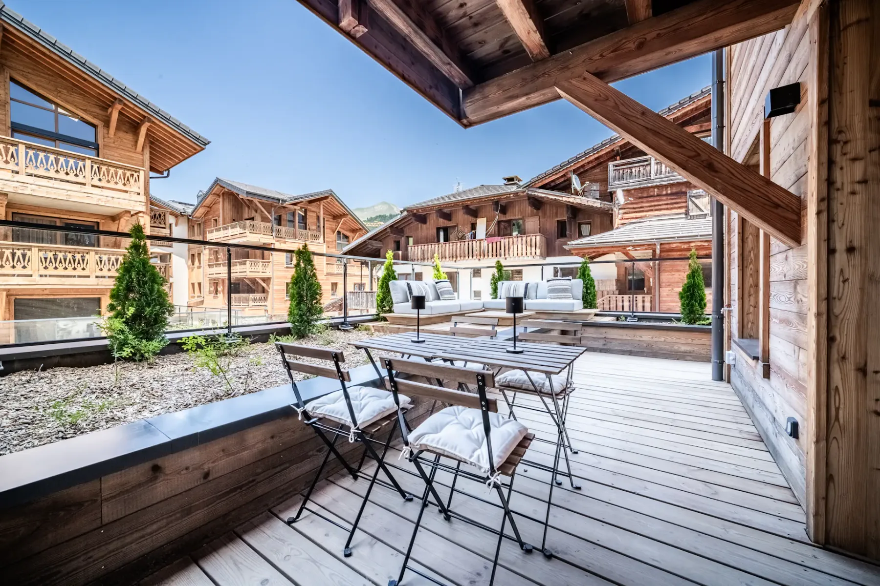 Outdoor wooden terrace with a table and cushioned chairs, overlooking an alpine-style village with wooden buildings under a clear blue sky.