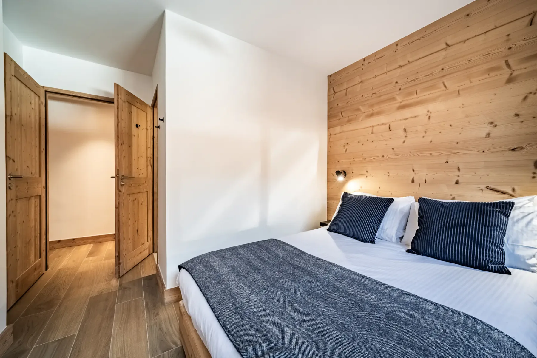 A cozy bedroom with wooden walls, featuring a double bed with blue pillows and a gray blanket. A window with curtains lets in natural light, and a pendant lamp hangs from the ceiling.