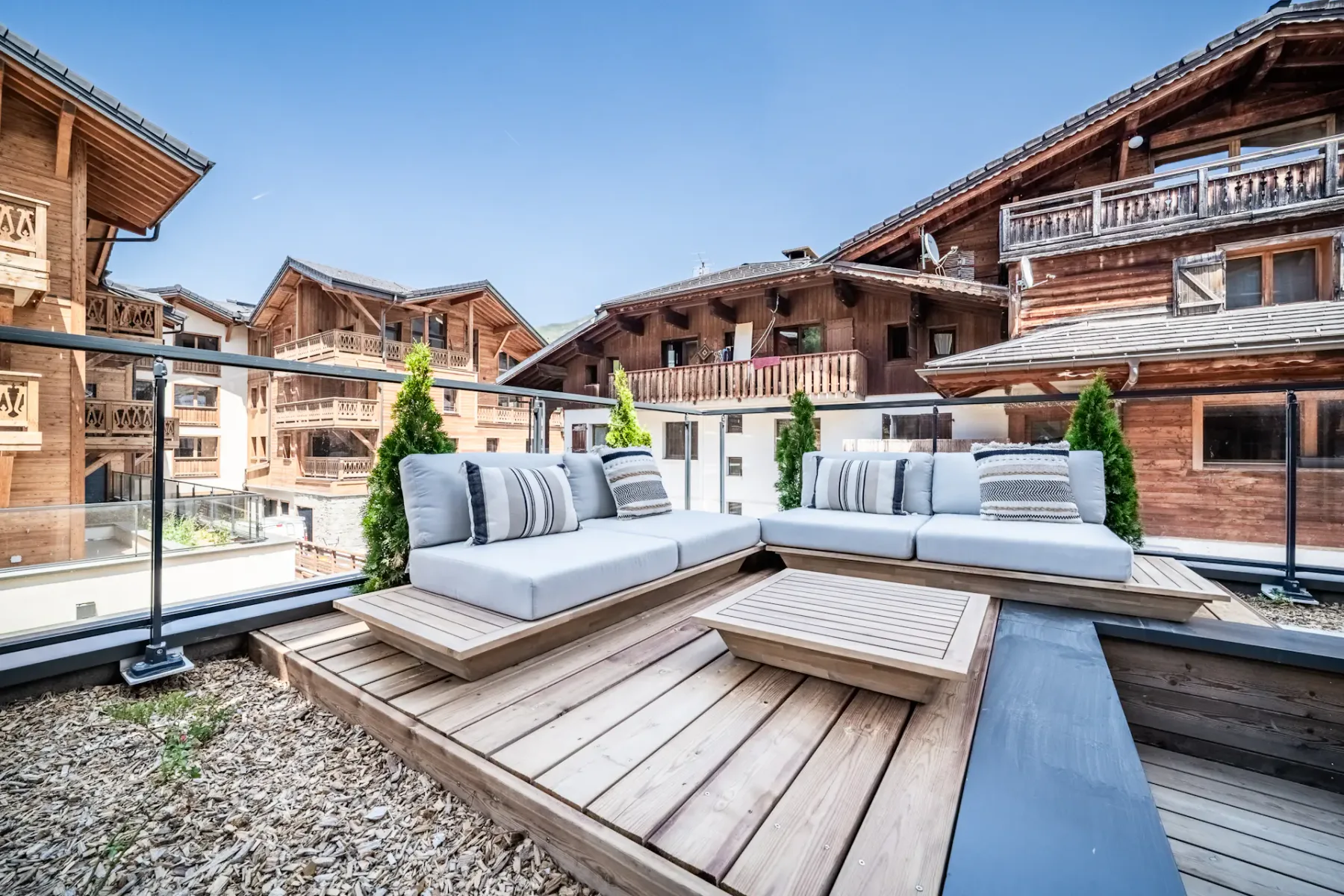 Outdoor wooden terrace with a table and cushioned chairs, overlooking an alpine-style village with wooden buildings under a clear blue sky.