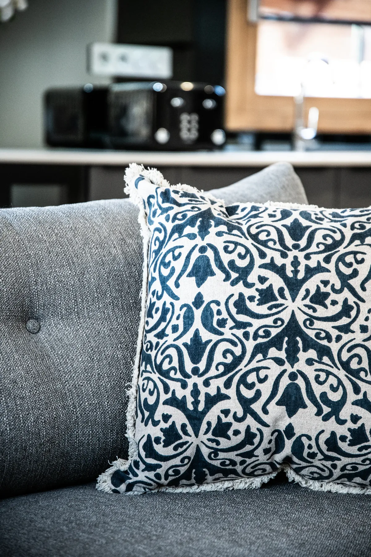 Patterned cushion on a gray sofa in a modern living room setting, with kitchen appliances visible in the background.