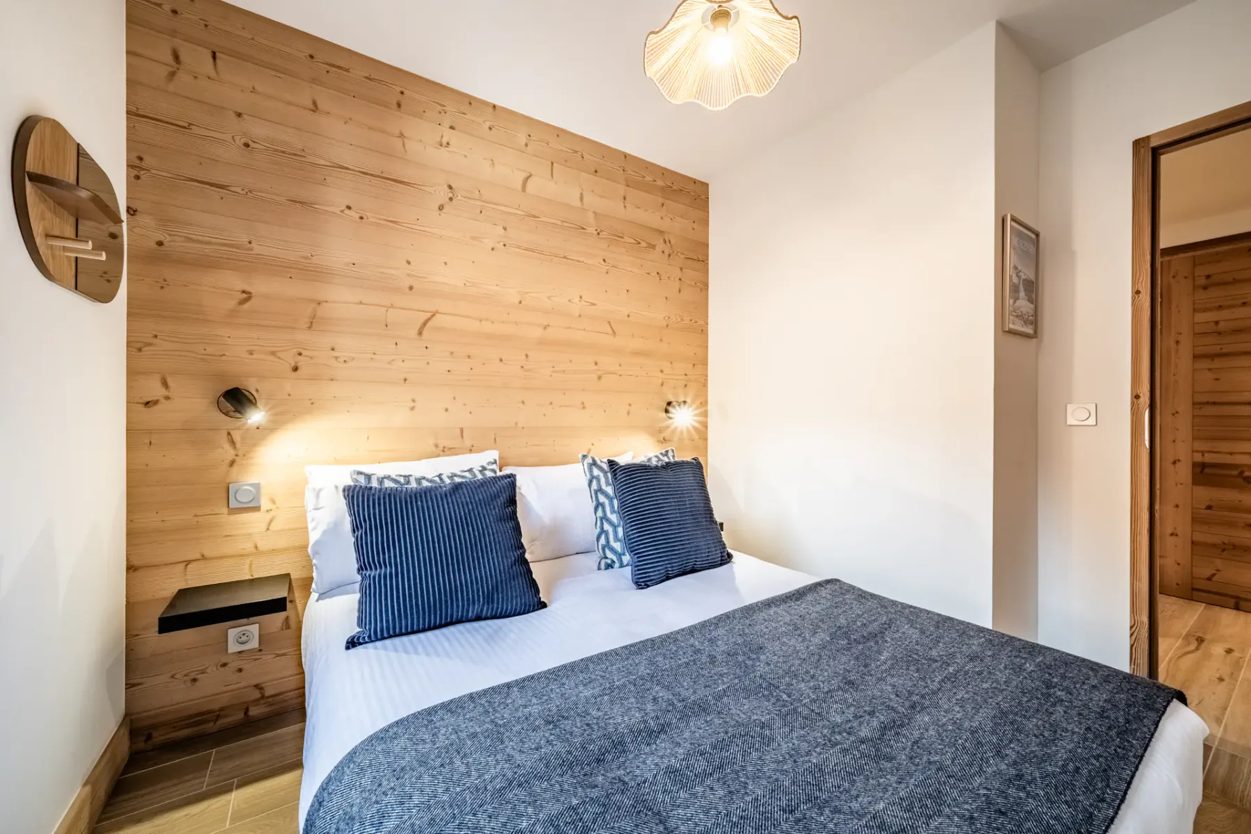 A cozy bedroom with a double bed, blue pillows, and a blanket. Wood-paneled wall with small light fixtures, a window, and clothes rack with hangers.