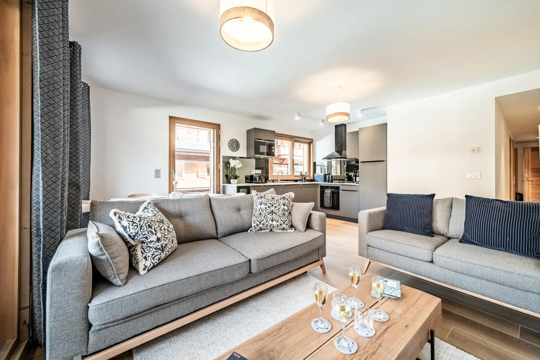 Modern living room with two beige sofas, a wooden coffee table, floor lamp, and wall-mounted TV. Glass doors open to a balcony with a view of neighboring buildings. Wood flooring and decor.