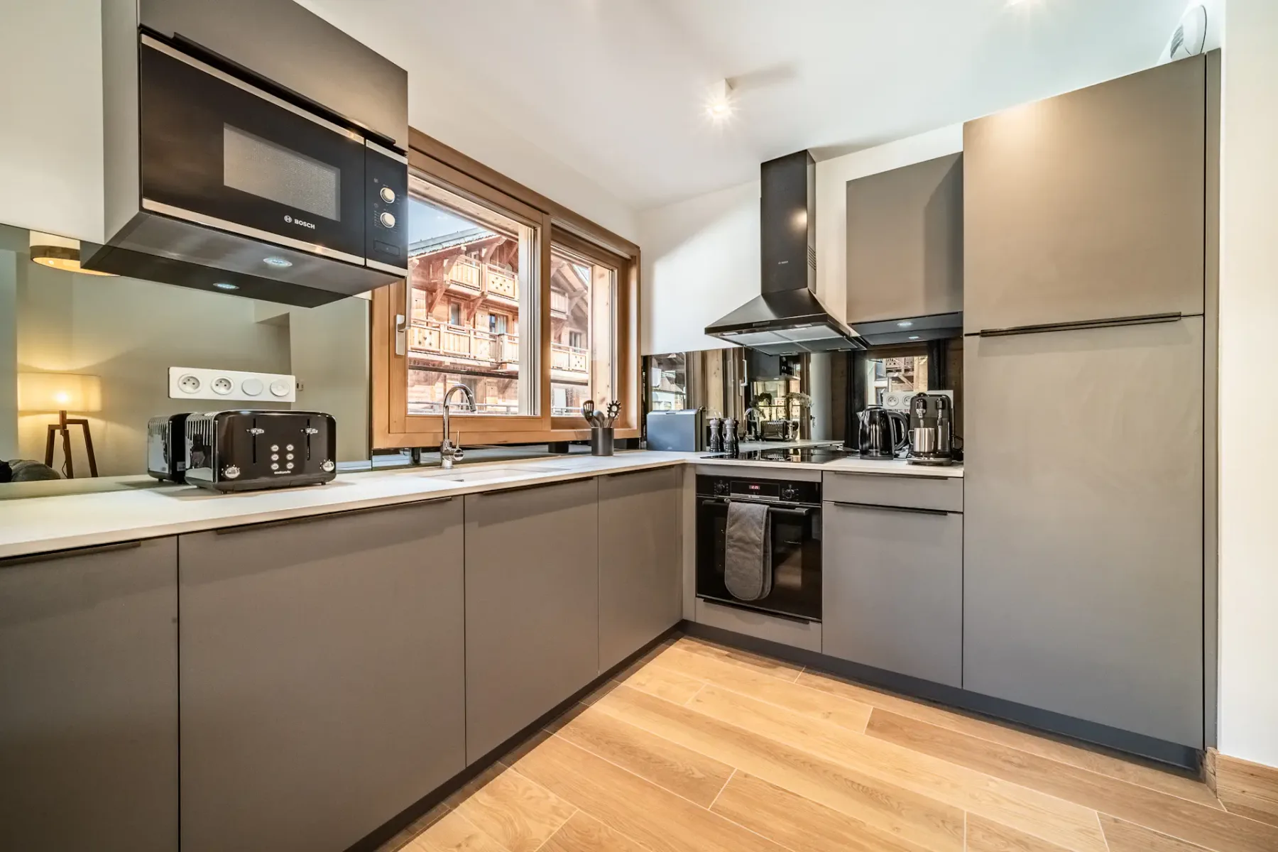Modern kitchen and dining area with gray cabinetry, wooden floors, a small dining table with chairs, a sofa, and large windows with views of wooden buildings outside.