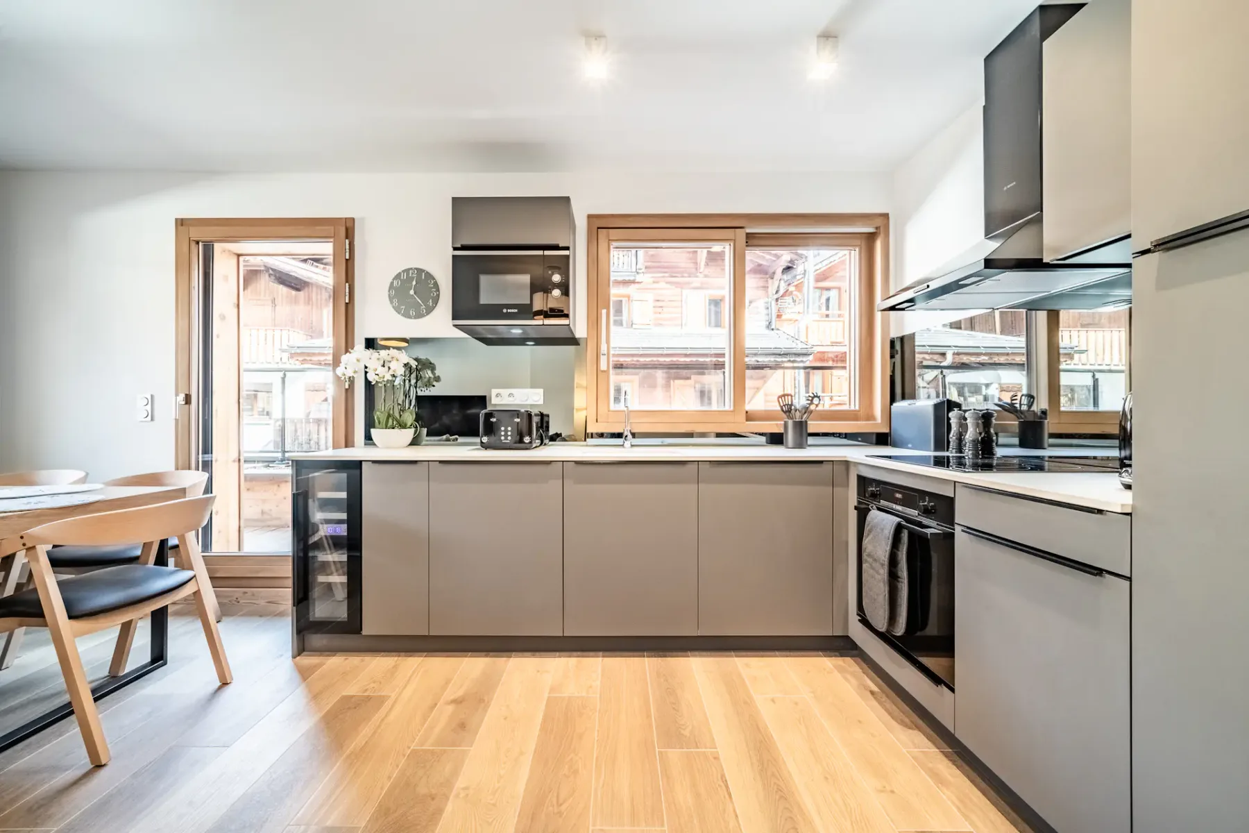 Modern kitchen and dining area with gray cabinetry, wooden floors, a small dining table with chairs, a sofa, and large windows with views of wooden buildings outside.