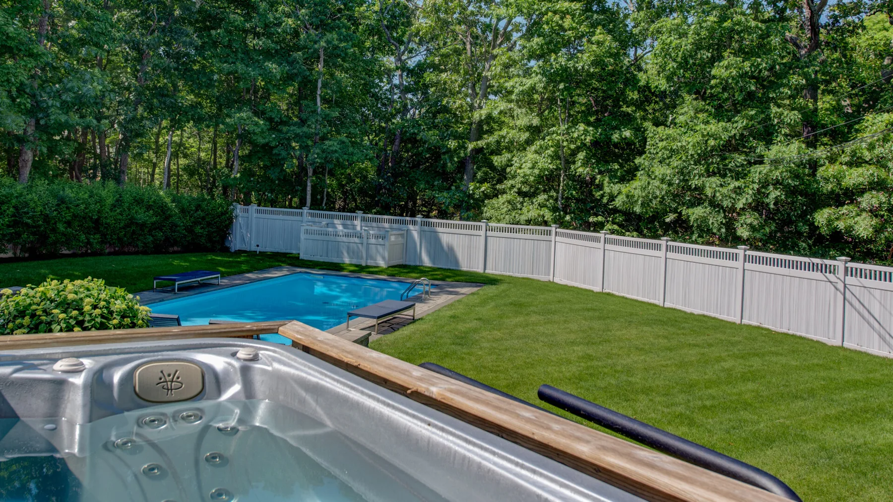 A backyard pool and hot tub sit beside a green lawn and trees.