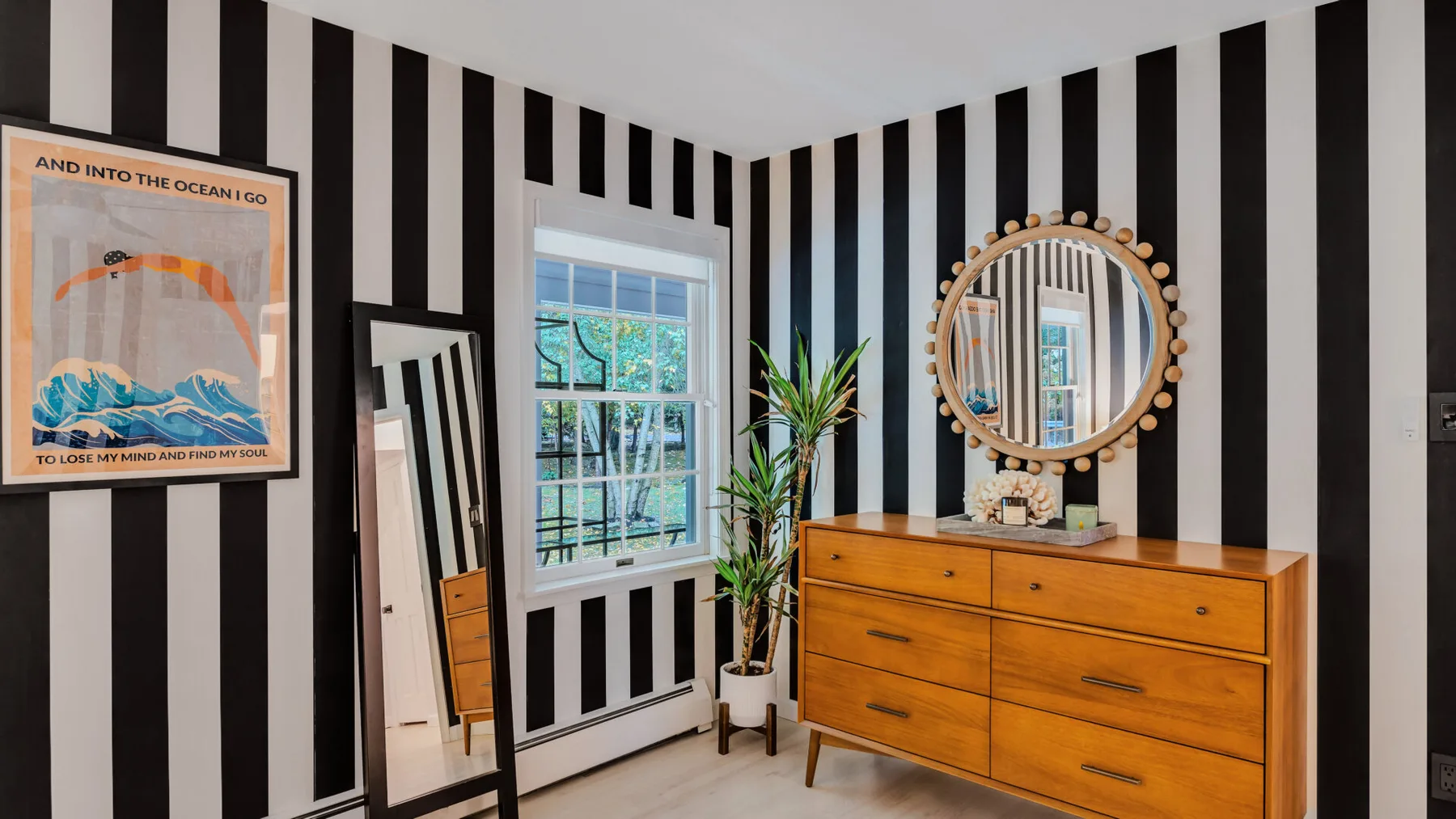 A bedroom with a wooden dresser, round mirror, and black-and-white striped walls.