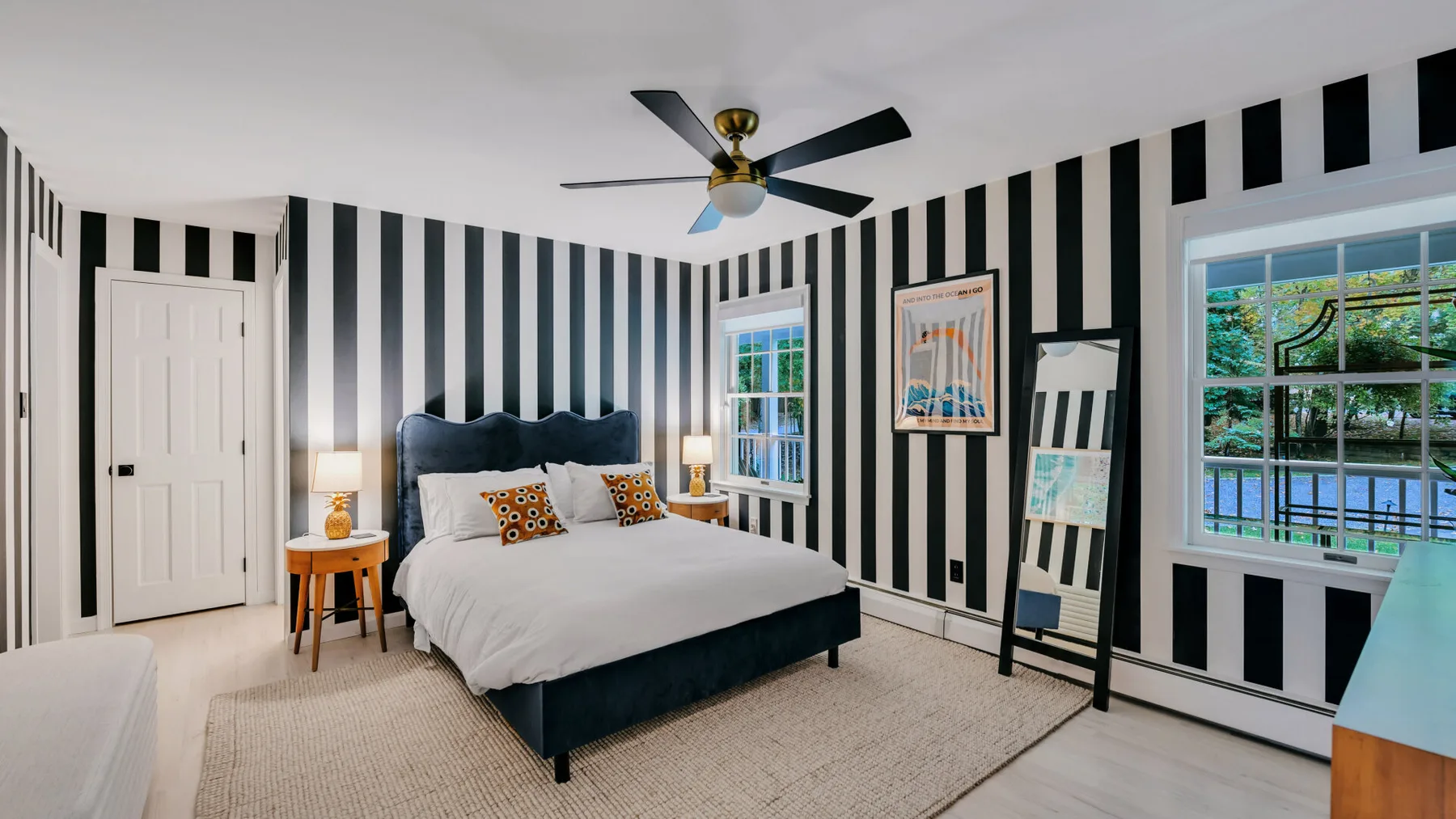 A bedroom with a bed, striped walls, and windows overlooking greenery.