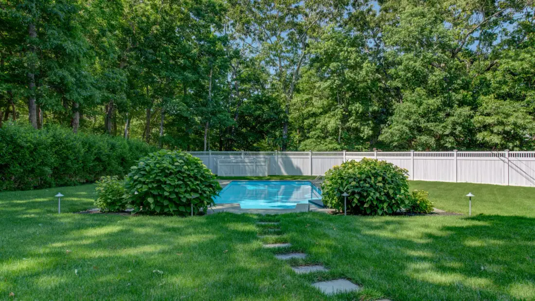 A backyard lawn leads to a swimming pool bordered by shrubs and trees.