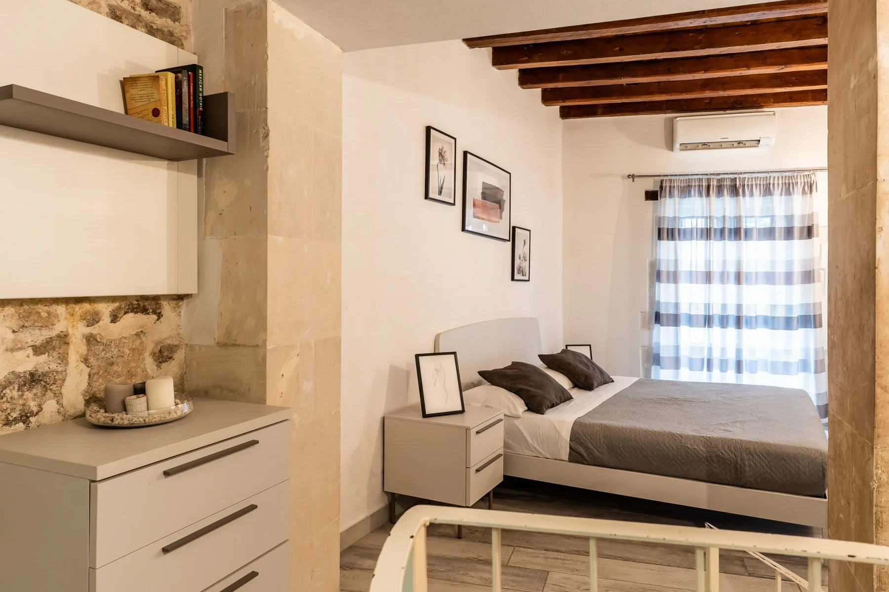 Un dormitorio con una cama, una mesita de noche y cortinas a rayas bajo vigas de madera vistas.