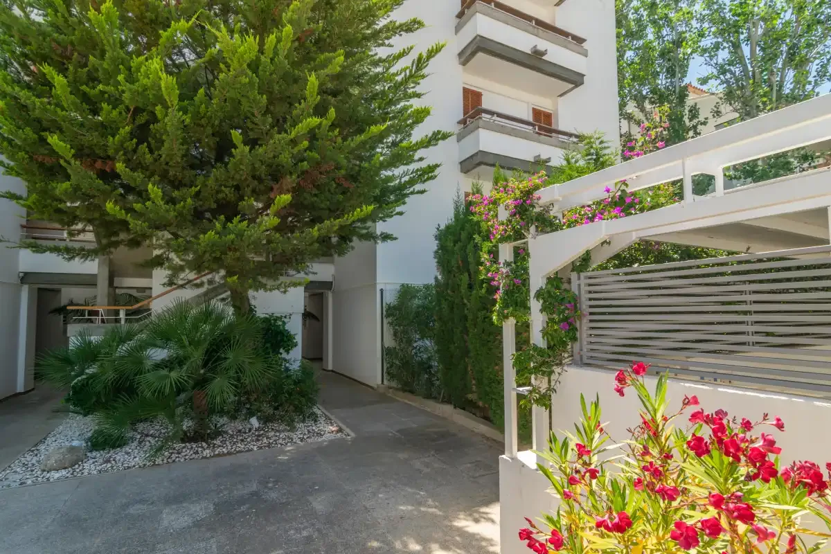 An apartment building entrance beside a paved walkway, trees, and flowering plants.