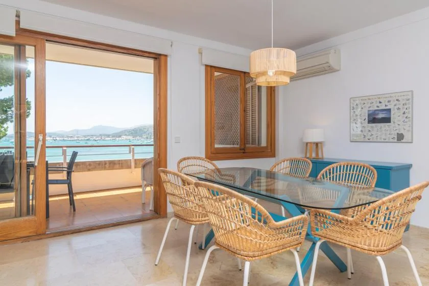 A bright dining area with a glass table and woven seating beside open balcony doors.