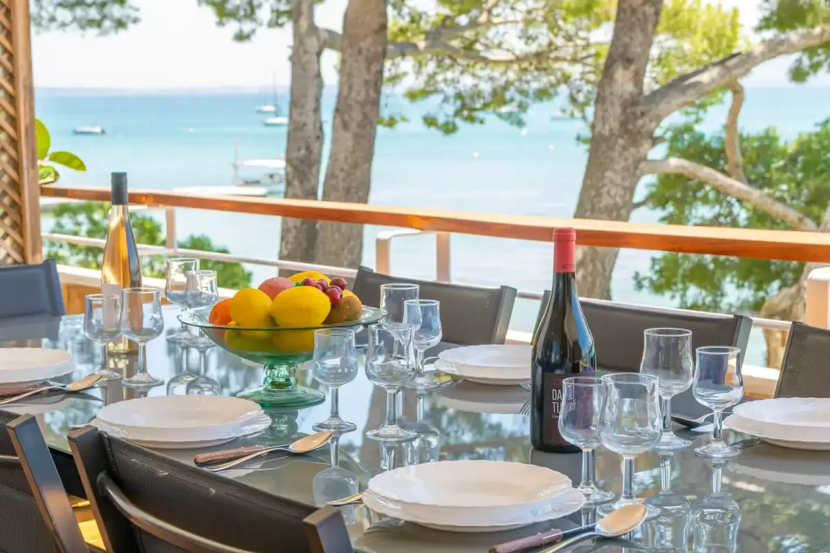 A dining table set with dishes and glasses overlooks the sea through trees.