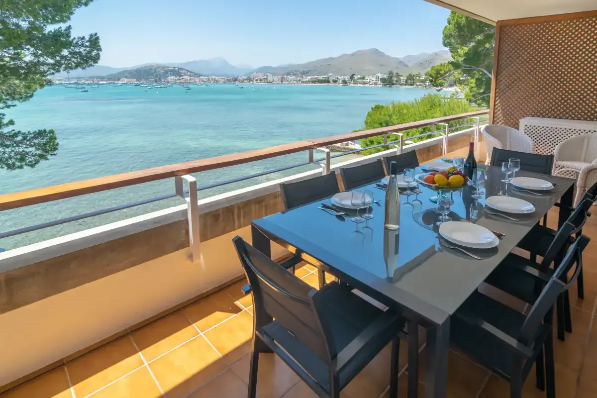 A furnished balcony dining area overlooks a turquoise bay and distant mountains.