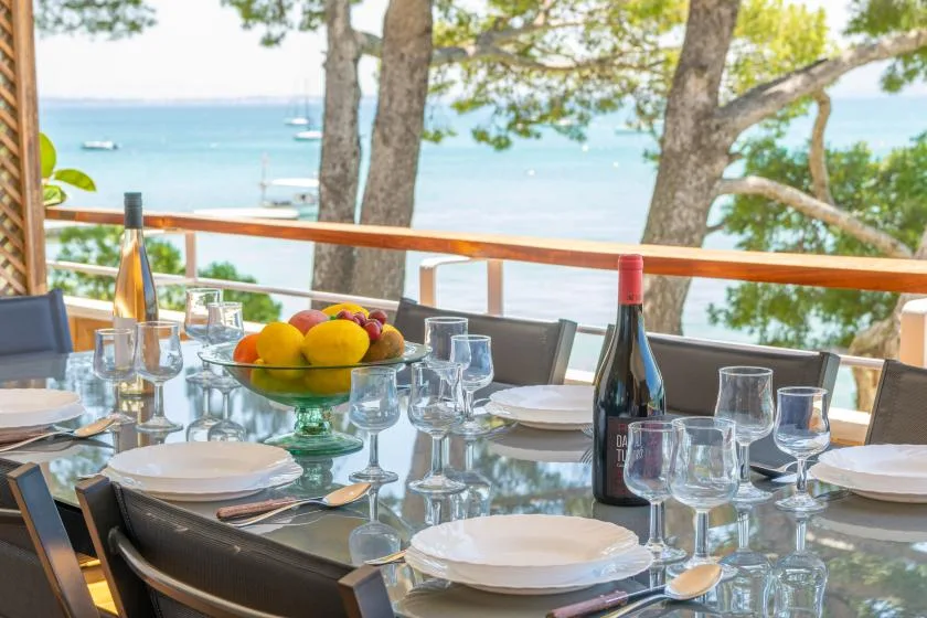 A dining table is set with glassware and fruit beside a waterfront view.