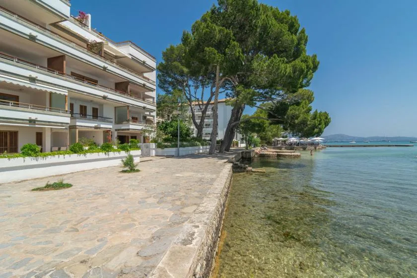 A paved waterfront promenade runs beside a white building and calm, clear water.