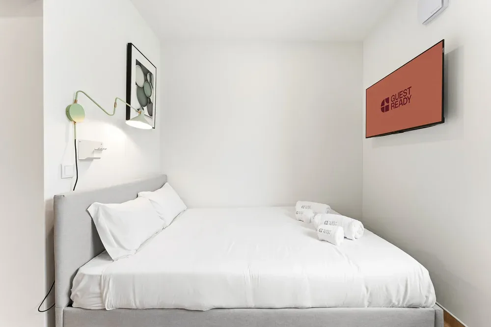 A bright bedroom with a neatly made bed and a wall-mounted screen.