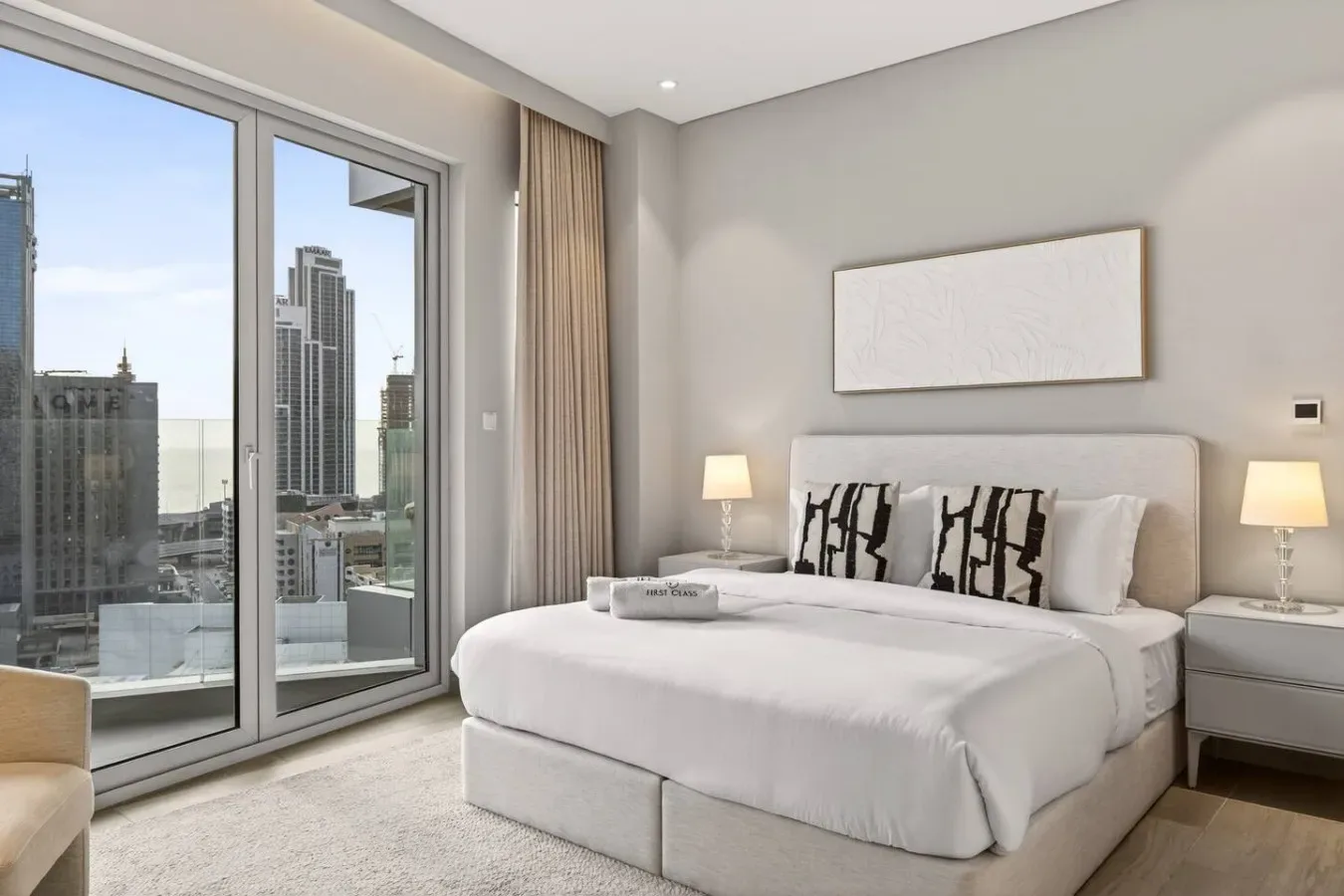 A bright bedroom with a neatly made bed and large windows overlooking city buildings.