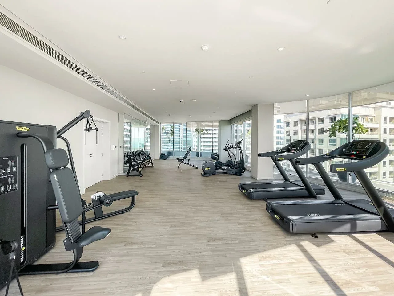 Un gimnasio luminoso con cintas de correr y equipo de ejercicio junto a grandes ventanas.