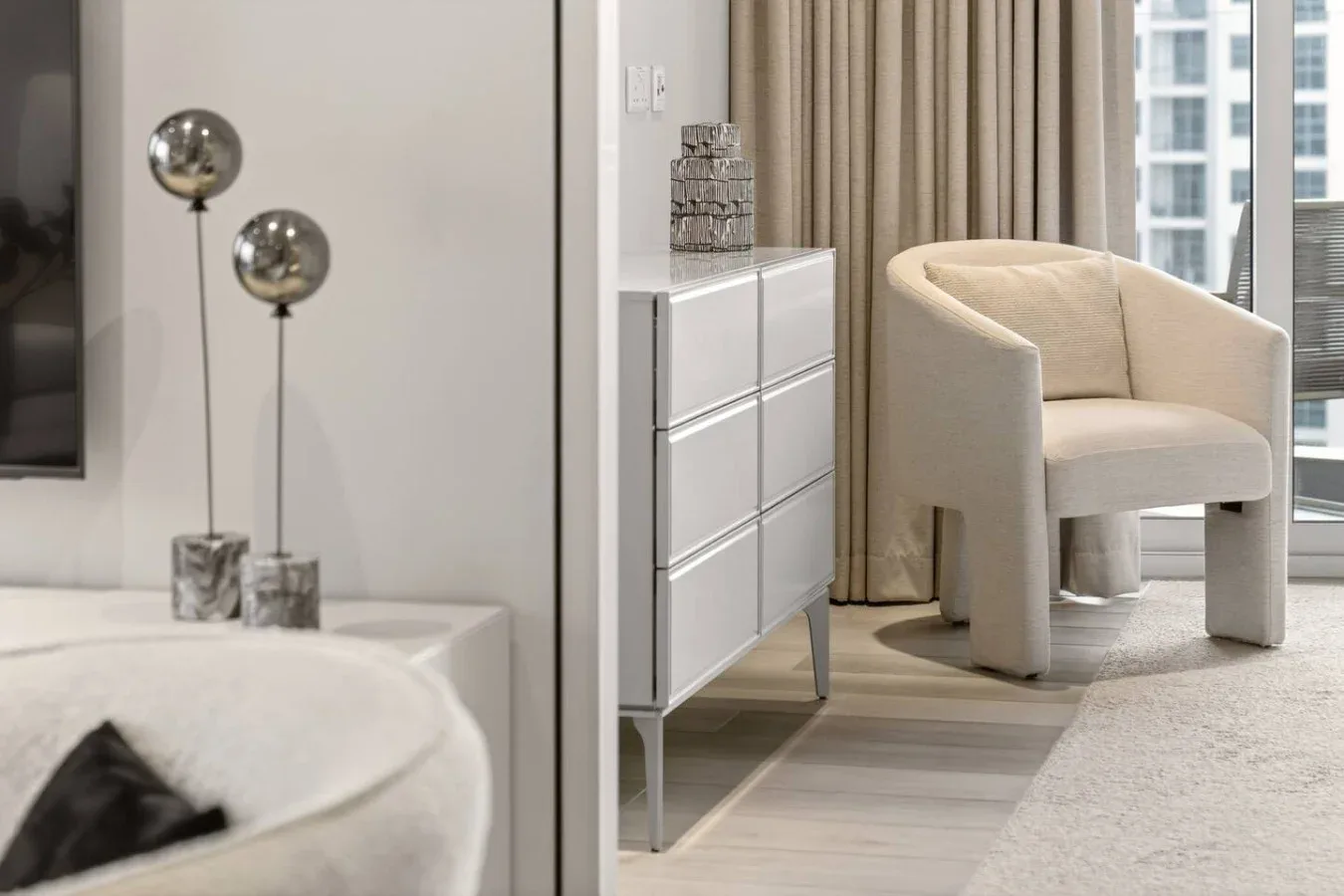 Un rincón de dormitorio luminoso con un sillón junto a una cómoda y una ventana.