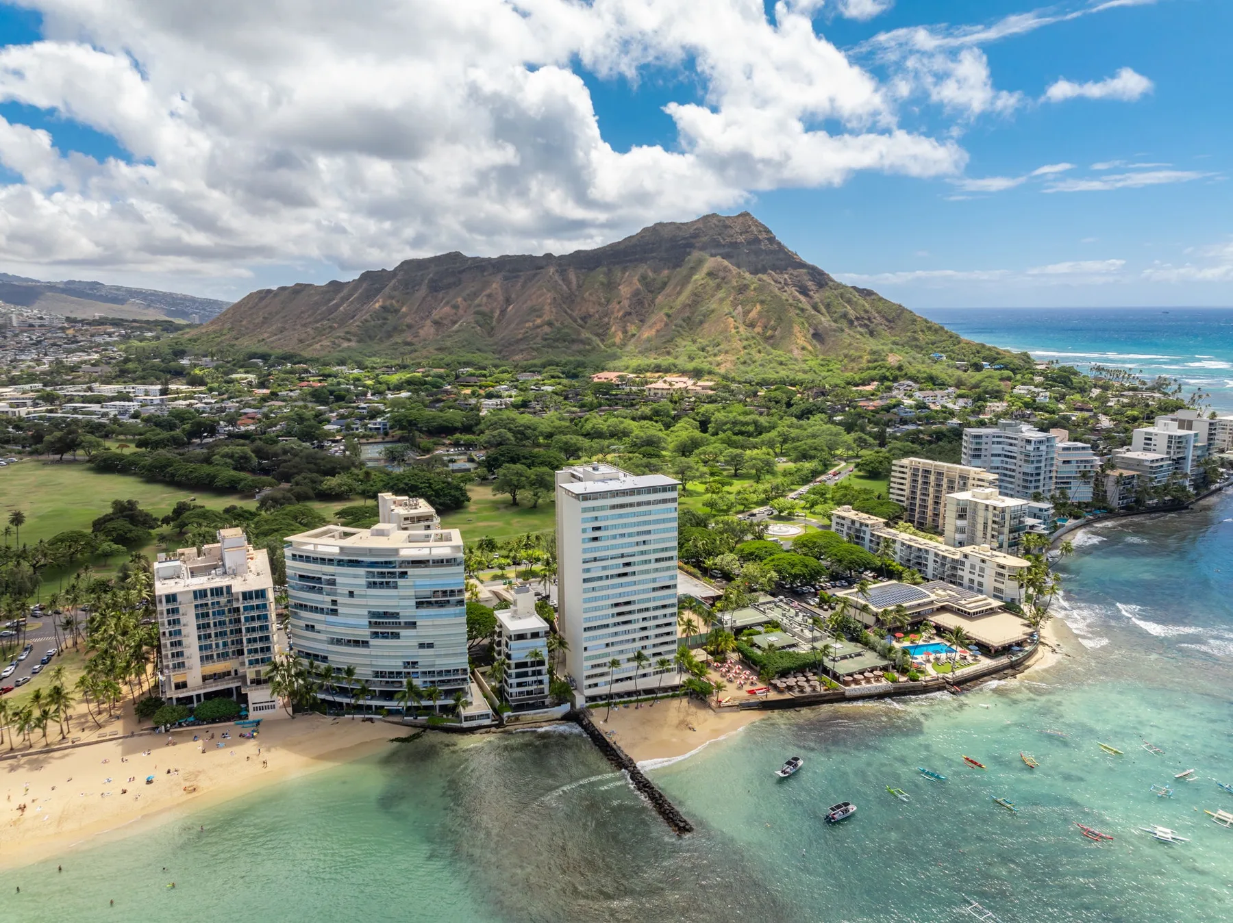 Aerial coastal view of a beach, waterfront buildings, and a green mountain.