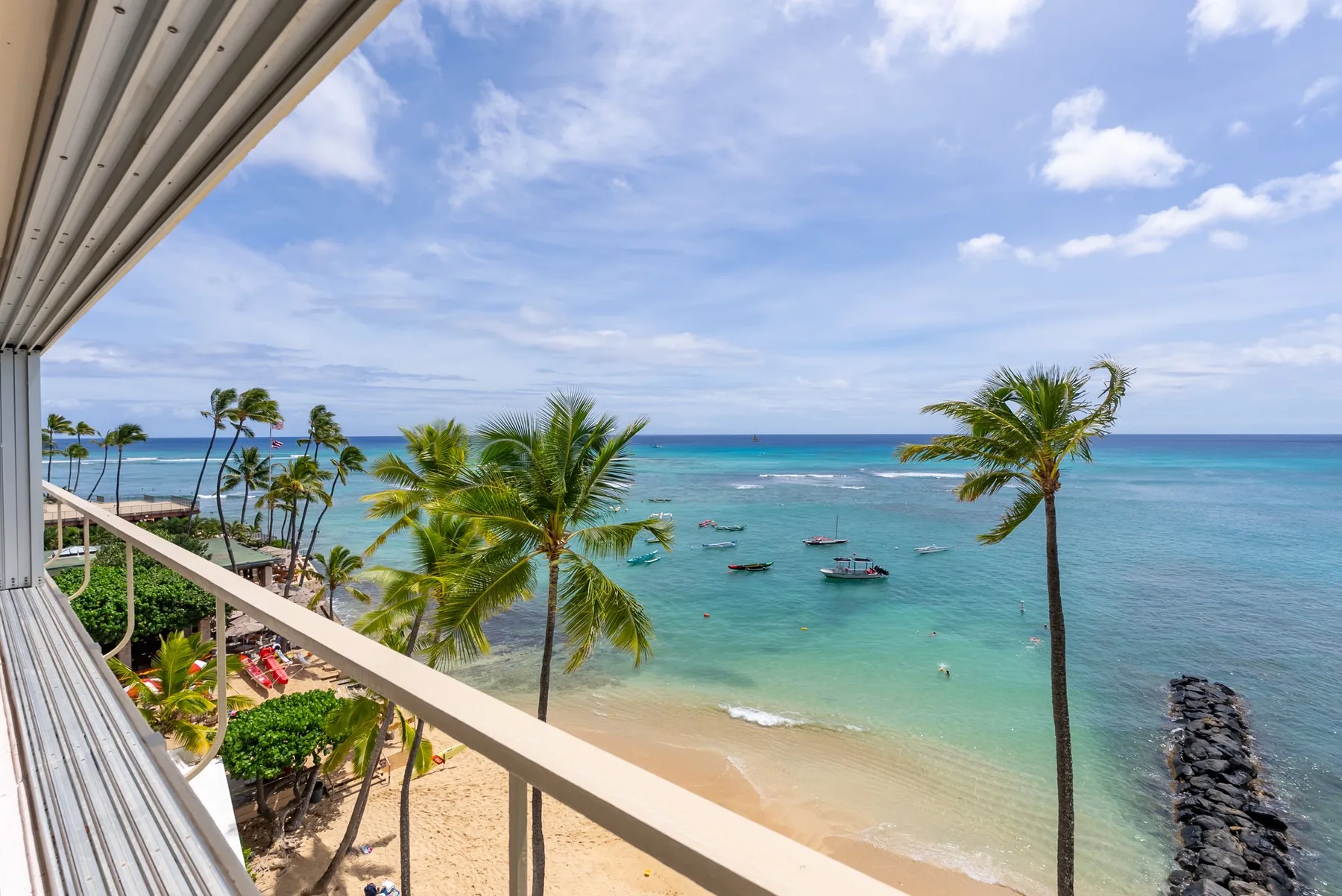 A beachfront balcony overlooks turquoise water, palm trees, and boats.