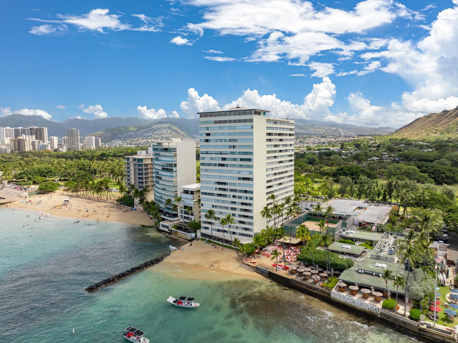 A beachfront hotel building overlooks a sandy shore and calm blue water.