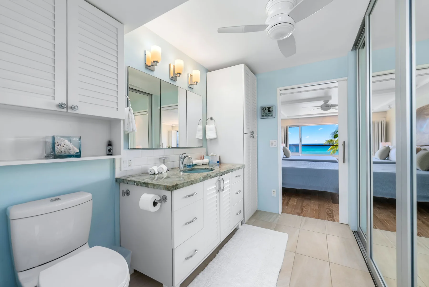 A bright bathroom with a vanity, toilet, and doorway to a bedroom.