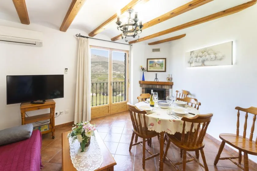 Comedor con la mesa puesta, un televisor y puertas que dan a un balcón.