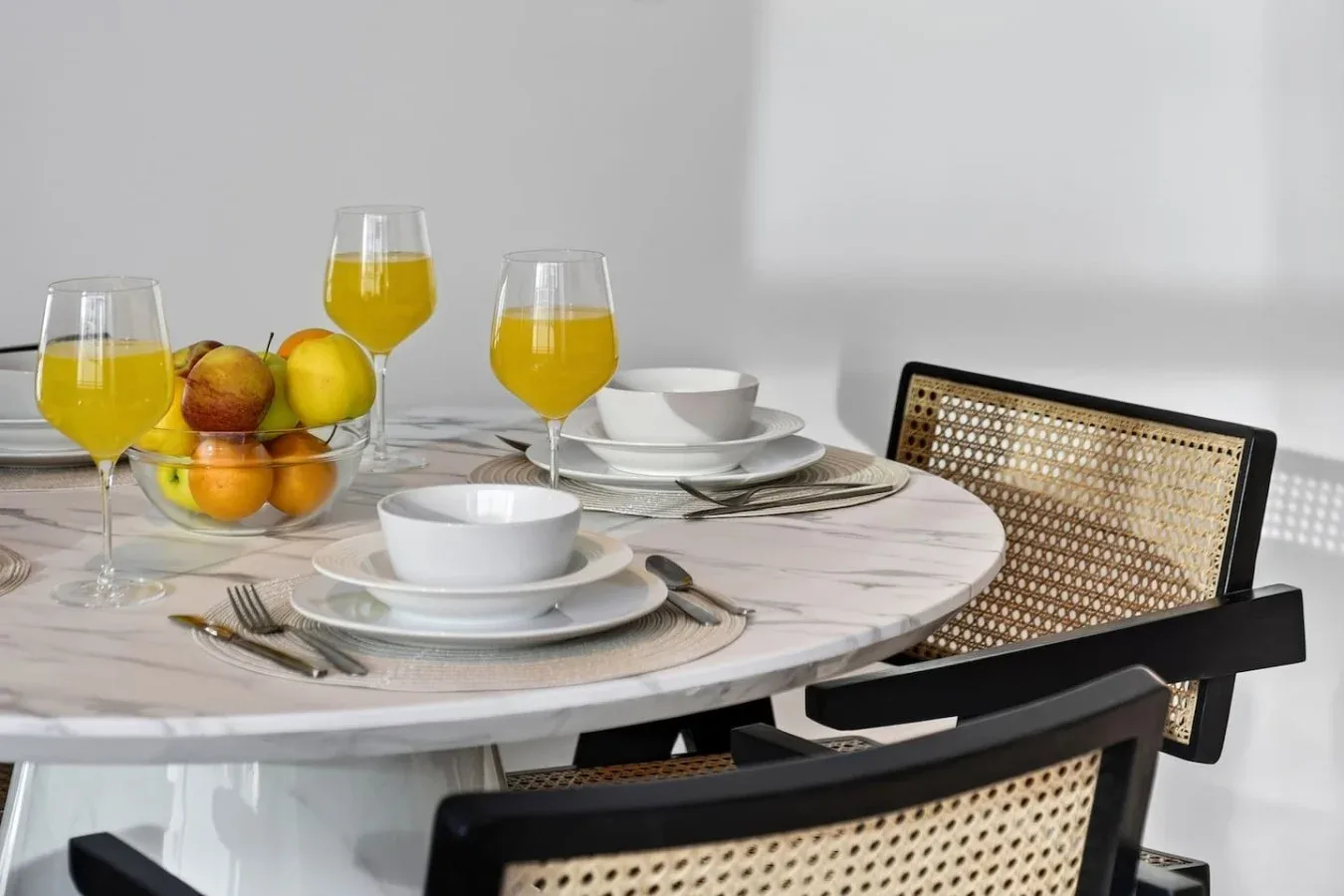 A dining table is set with plates, glasses of juice, and a bowl of fruit.